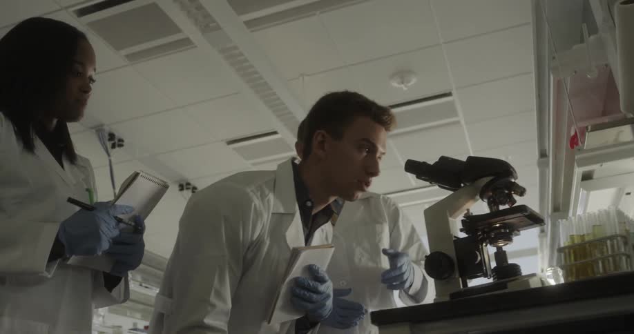 A teacher nods and instructs as two lab students take notes then one bends and looks through a microscope