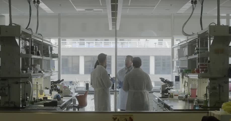 Scientists in lab coats and wearing gloves take notes while another explains in a lab