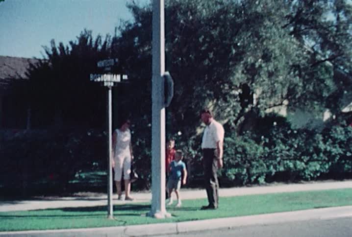 Old footage of a man and women standing with two boys who are holding lunch bags on a corner