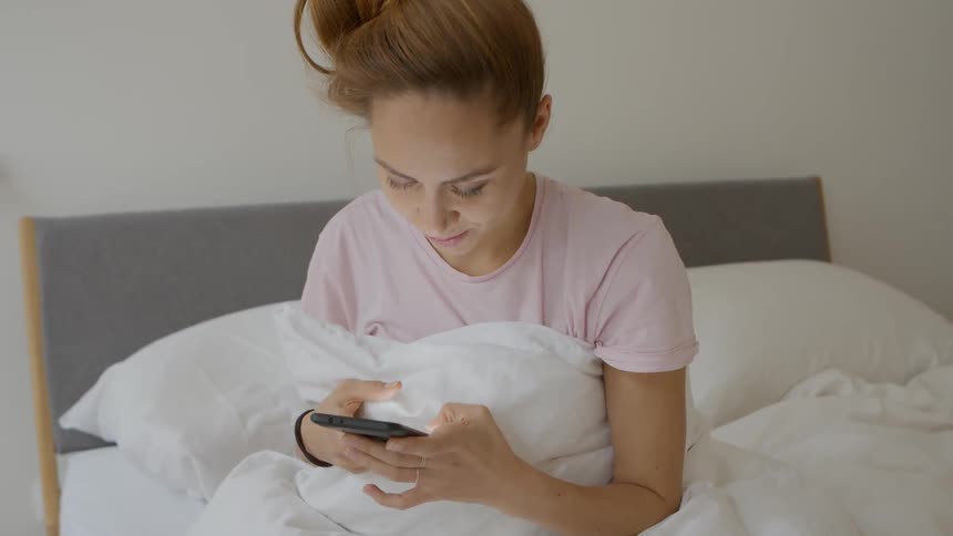 A woman types on her smartphone while sitting in bed