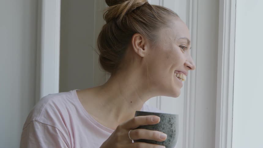 A woman is smiling as she holds a mug by the window