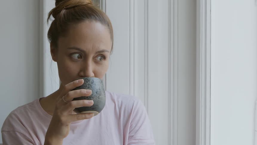 A woman standing at a window holding a teacup drinking tea