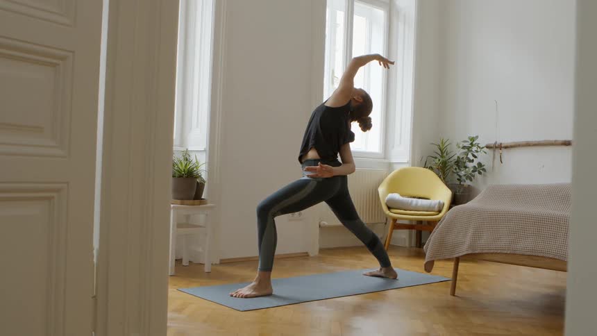 Female practicing yoga on a yoga mat placed on the hard wood floors of her home bedroom as the sunlight shines through the windows