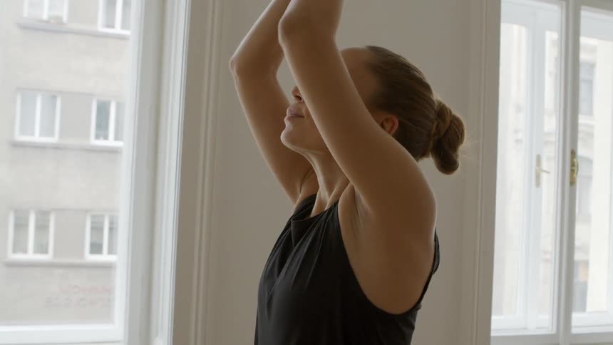 A woman stretches her arms high in the air and brings her hands down folded in front of her chest