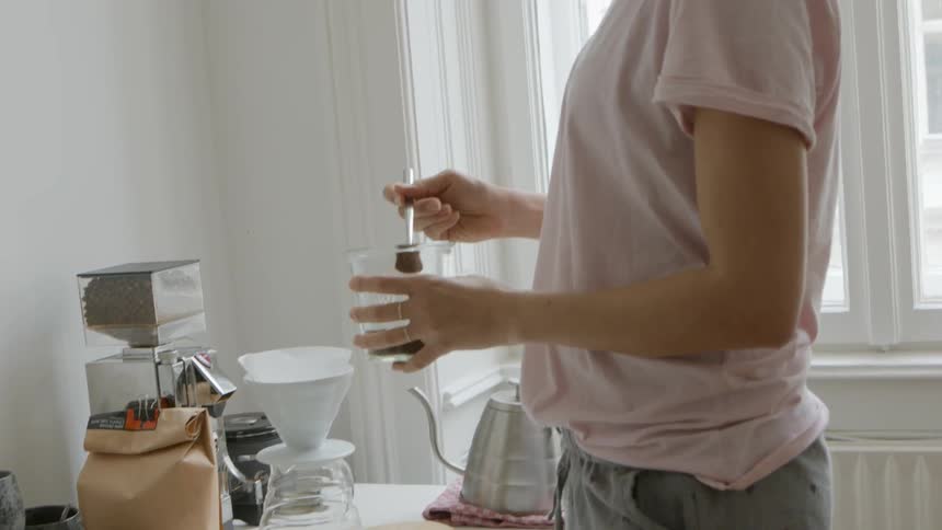 Female picks up a spoon with a long handle and scoops coffee grounds from a glass container into a coffee filter