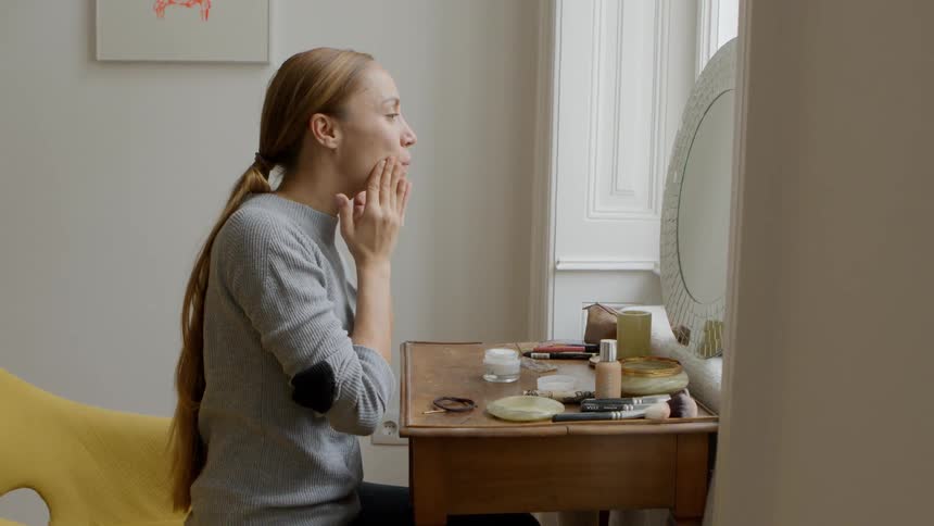 A woman rubs moisturizer all over her face while sitting at her vanity