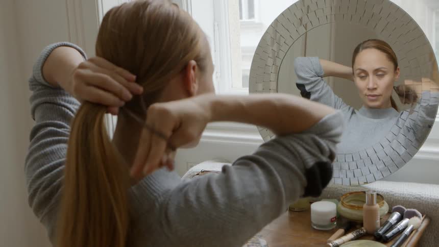 A woman sits in front of her vanity and ties her hair back in a low ponytail