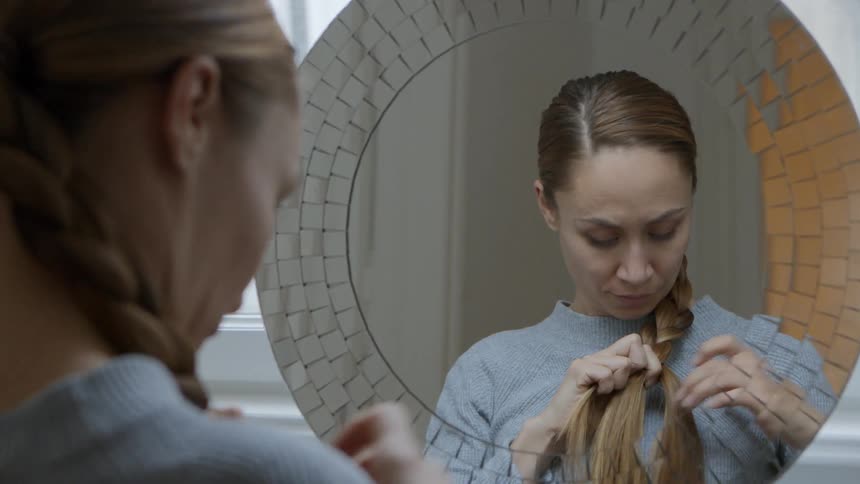A woman braids her hair while staring into a mirror