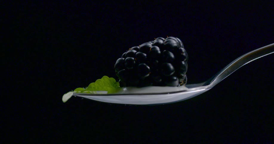 A blackberry sits on a spoon with yogurt and a mint leaf