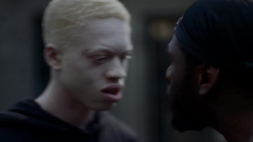 A shirtless black man yells at and pushes an albino teenager