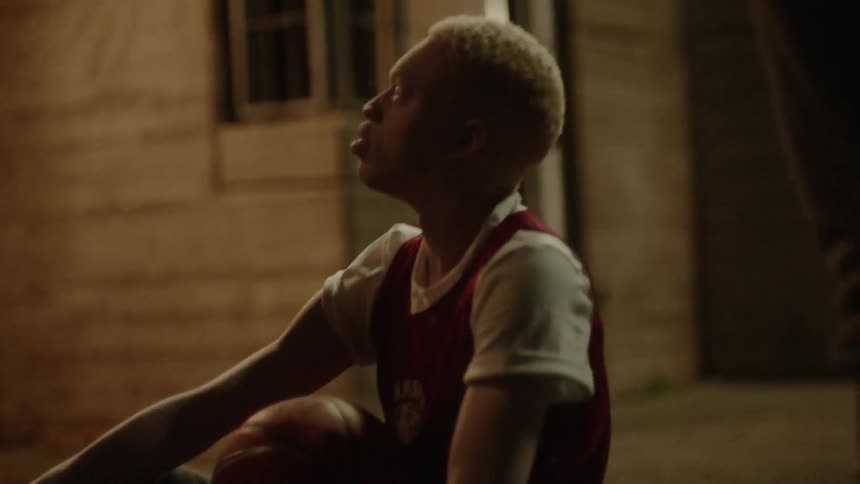 A young man is sitting on the ground in a courtyard with a basketball on his lap as two other men look down on him