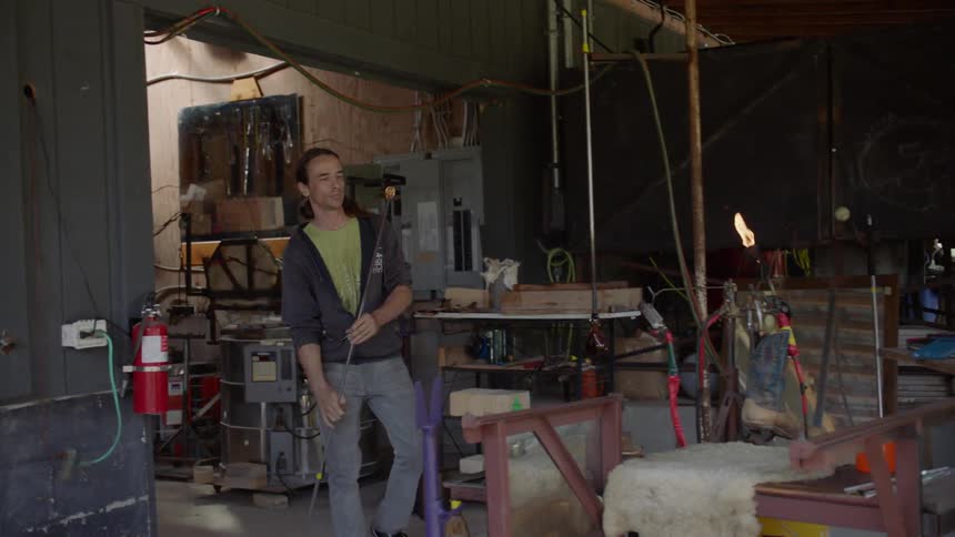 Man blows on molten glass blob topping metal pole