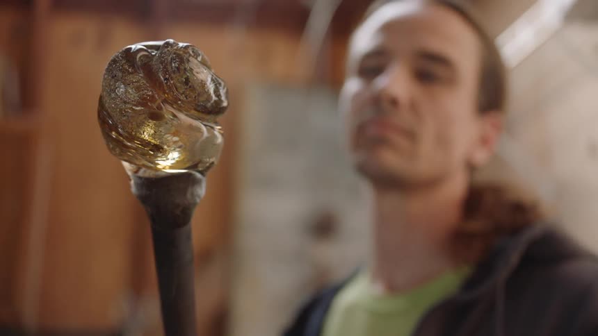 A glass blower stares at a piece of molten glass on a pole