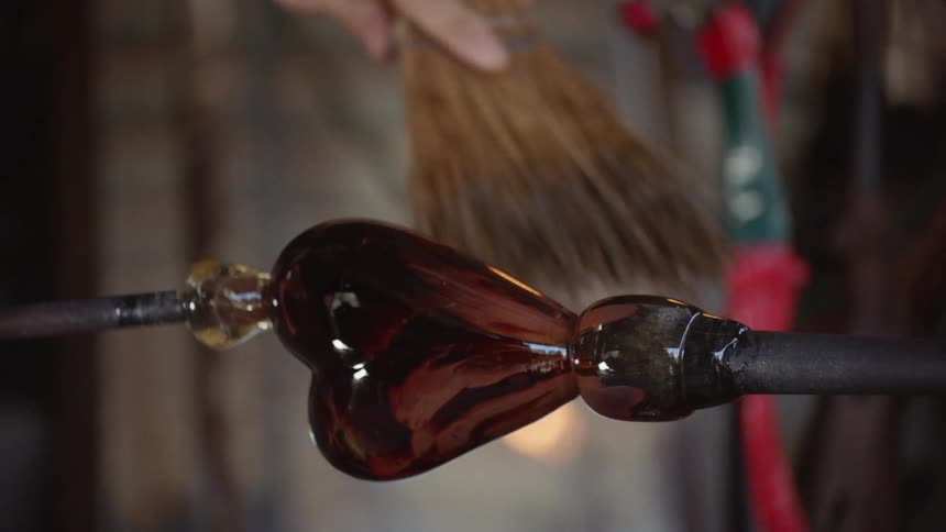 A glass blower brushes a piece of glass with a broom