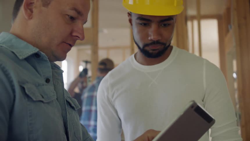 A man holds a notepad and talks to a construction worker wearing a hardhat