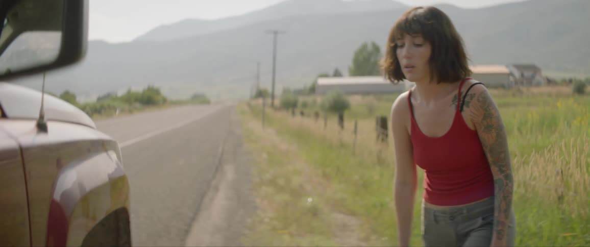 Female who has a flat tire on the side of a deserted road looks at her car with a sigh and moves forward to put on a spare tire