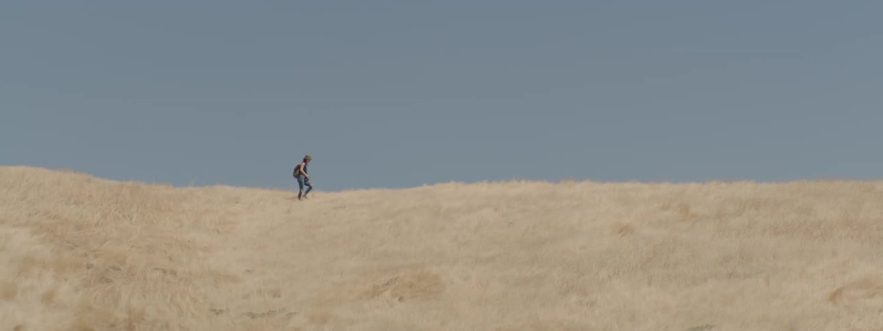 A young woman walks across the top of a grassy hill
