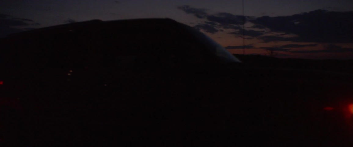 A car with its headlights on drives in a rural field at dusk