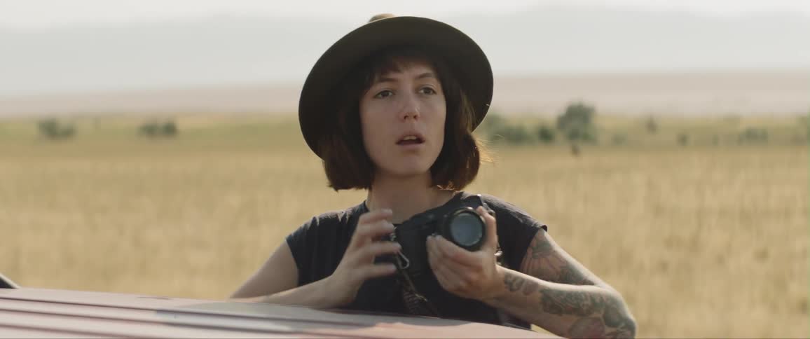 A woman pops up from inside a car and leans forward holding a camera looking around the grassy plain