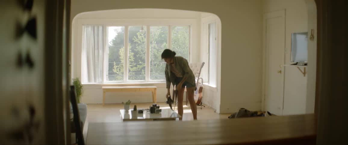 A woman walks into her home and puts her bags on the floor also putting a camera on the table and taking a jacket off