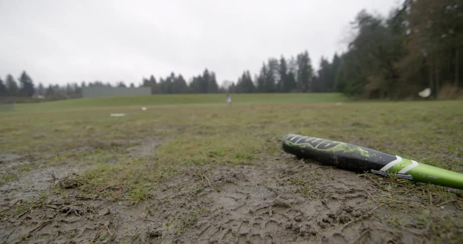 A baseball bat rests on the wet ground on a cloudy day while a boy runs around in a grass field in the background