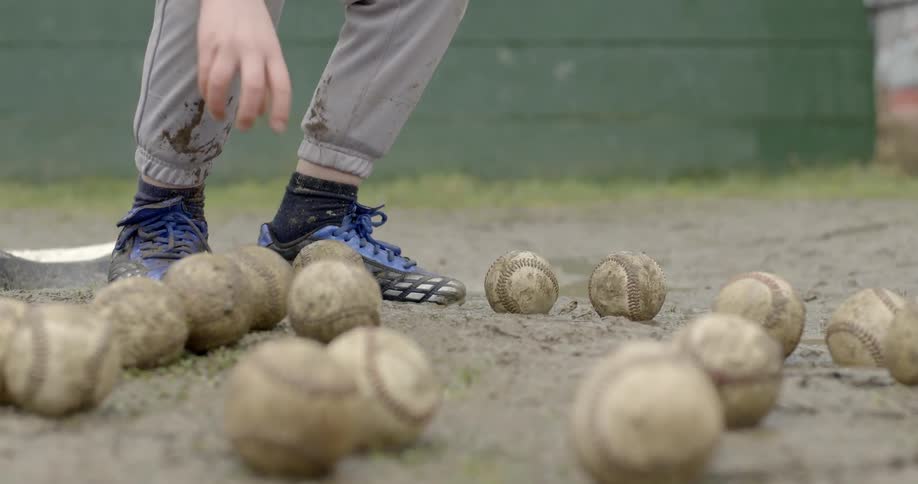 A boy walks towards a number of baseballs laying on the ground and picks one up and walks away