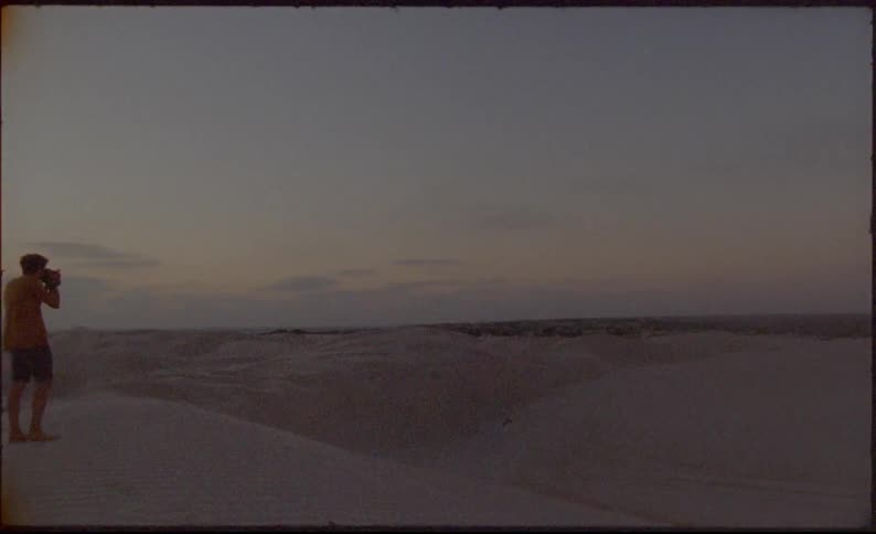 A person walking across sand dunes holds a camera up to their face