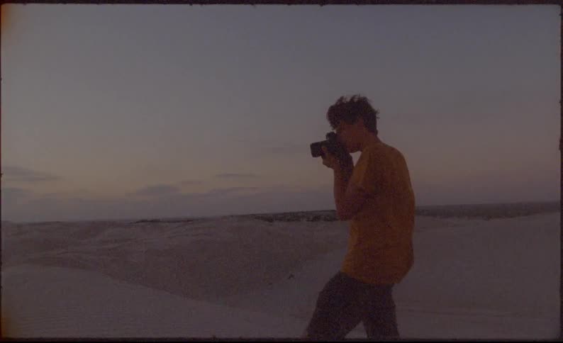 A man stands on a sand dune taking a photograph as the wind blows his tee shirt