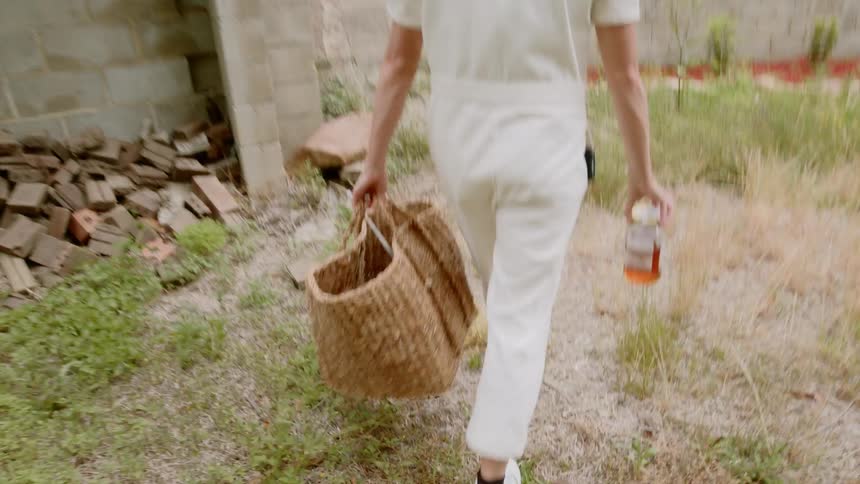 A beekeeper walks through her yard carrying a wicker bag and a jar of honey