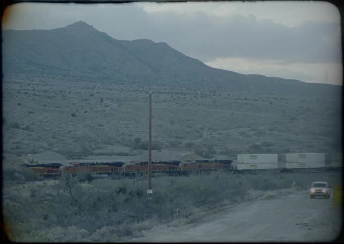 Train and car travel through scrub valley near mountain on a cloudy day