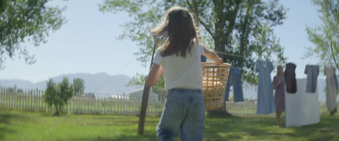 A girl carrying a laundry basket runs across the grassy yard to her mother near the clothesline