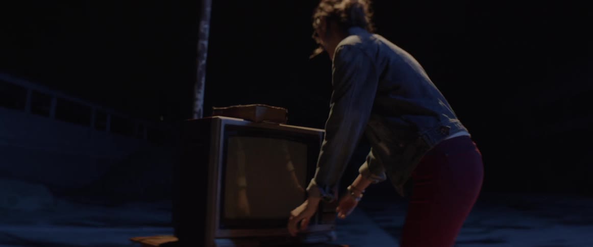 A woman walks through a dark parking lot and squats near and old television