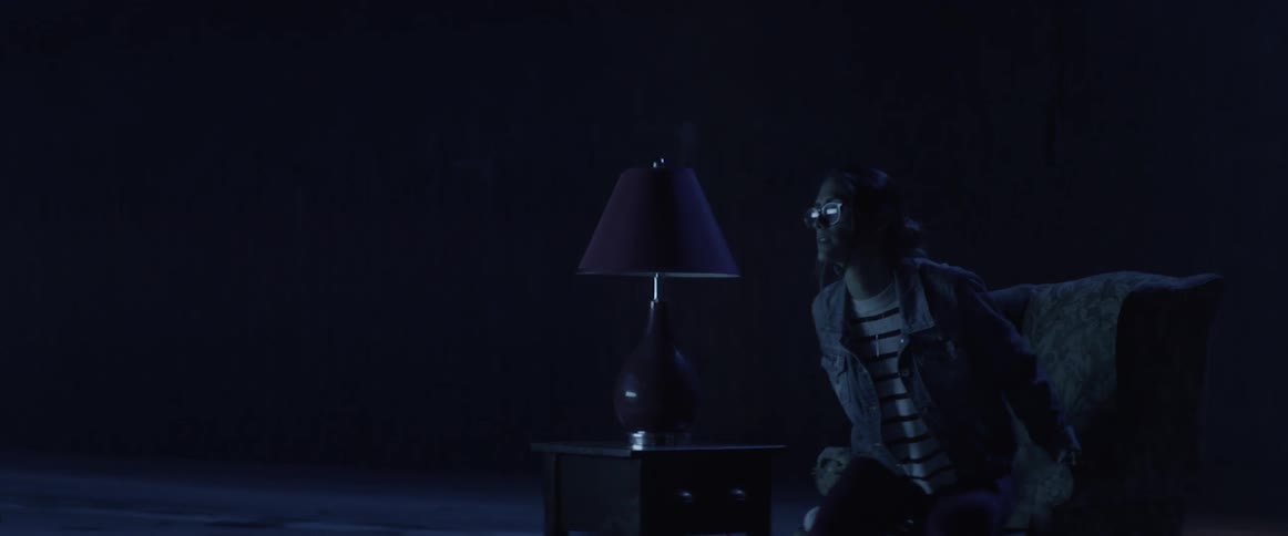 A woman sitting cross legged in a chair in a dark room before rising and walking away