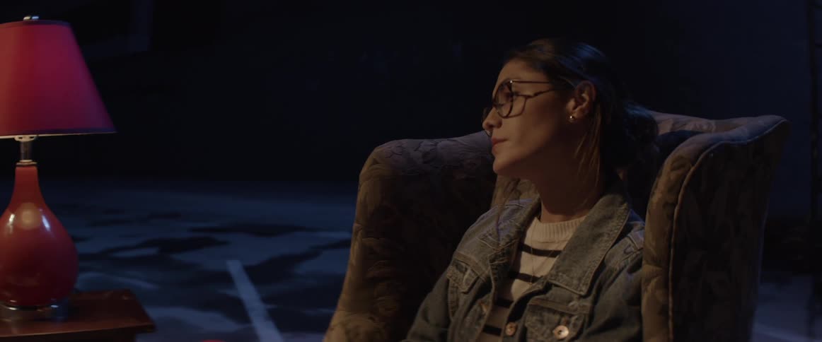 Woman sits in upholstered arm chair near lamp in dark room