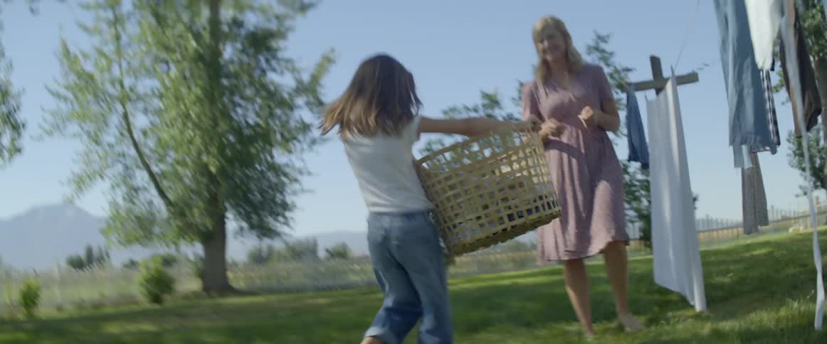 A little girl hurries to bring her mother a basket of clothes to place on a clothes line outdoors