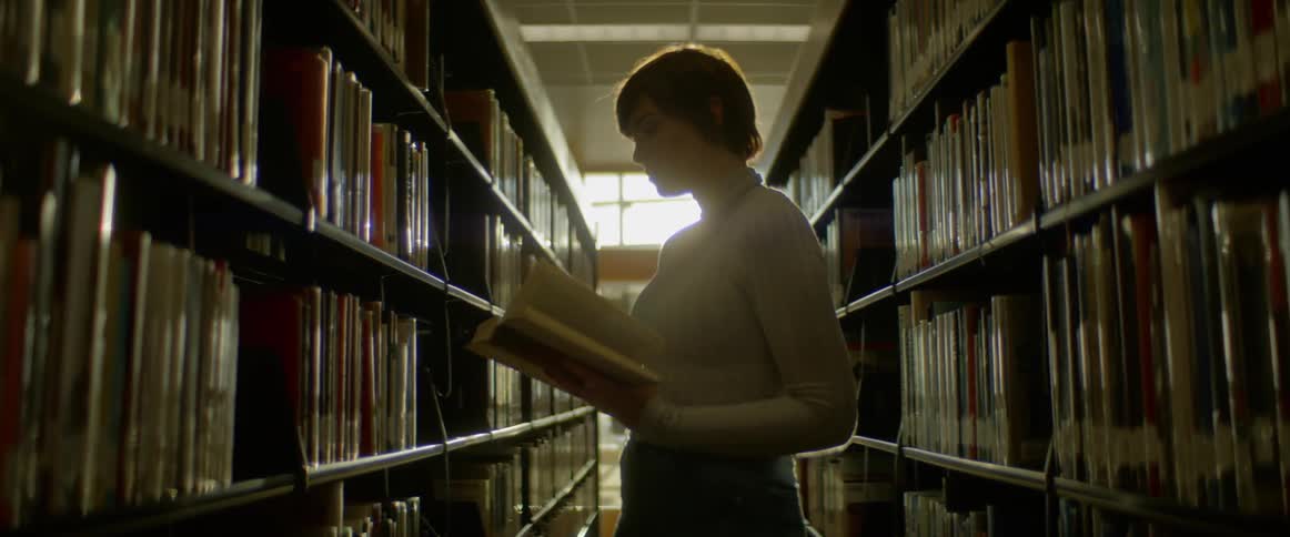A female with short hair is standing in the middle of two aisles of shelves in the library pulls a book off of the shelf and begins to turn the pages