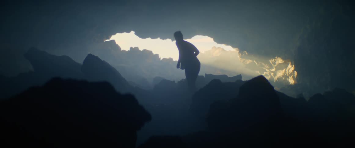 A woman walking through a cave before stopping to take a photograph of sunlight shining through an opening