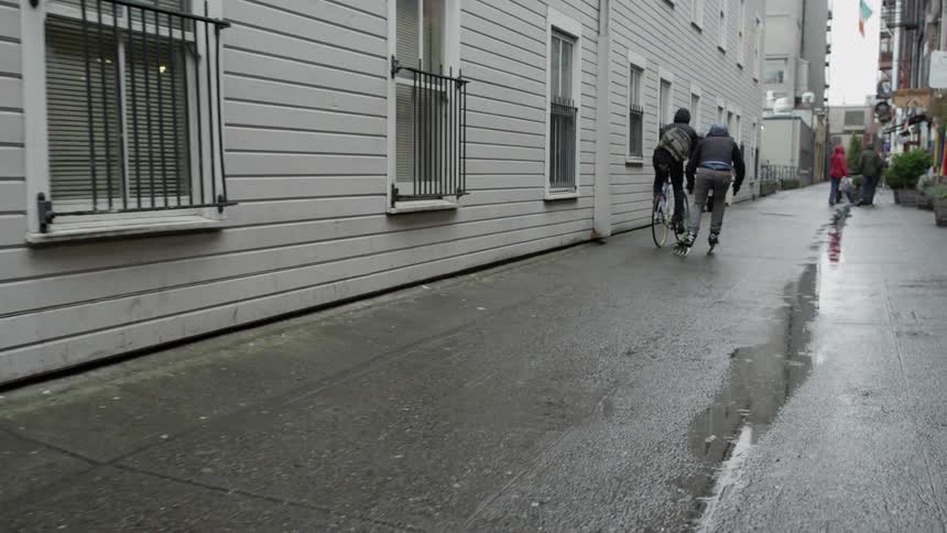 A man on skates carrying a camera rig skates beside a man riding a bike