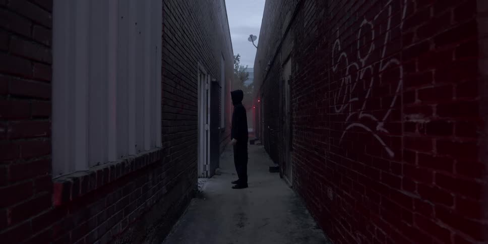 A man dressed in all black walks down an alley and goes through a door