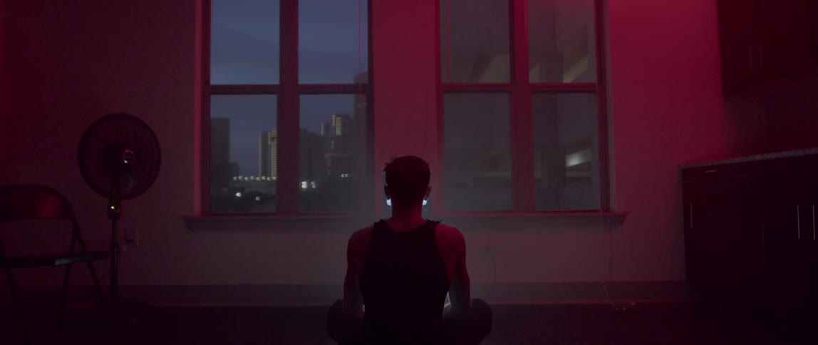 A man sits on the floor in a dark room as lights from a screen flash in front of him