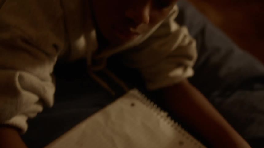 A black child writes in a notebook while laying on a bed