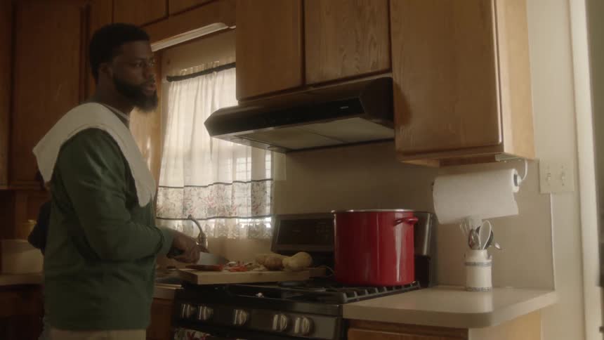 A boy walks past his dad standing at the stove chopping vegetables and out the door without pausing