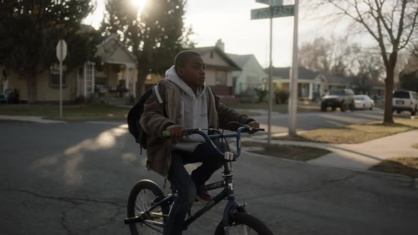 A boy pushes and then gets on his bicycle and rides down a neighborhood street