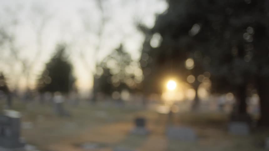 A boy wearing a hoodie and jacket standing in a cemetery at sunset bends over