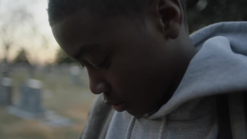 A young black male looks down contemplatively while standing in a cemetery as the sun sets