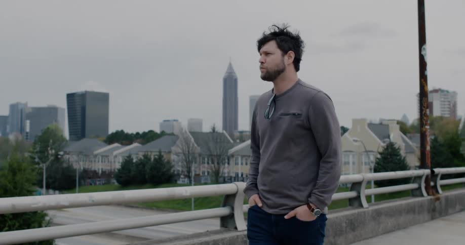 A young man with a beard slowly walks along an overpass above a city street near a residential area