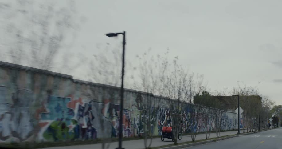 Several bike riders travel single file along a city sidewalk lined by a wall of graffiti on an cloudy afternoon