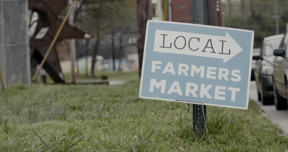 A small sign pointing in the direction of the local farmers market is swaying slightly in the grass next to cars parked along the side of the street