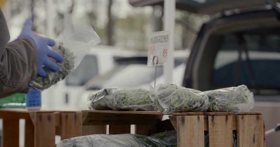 A person wearing gloves picks up a plastic bag of greens at an outdoor market and then puts it back