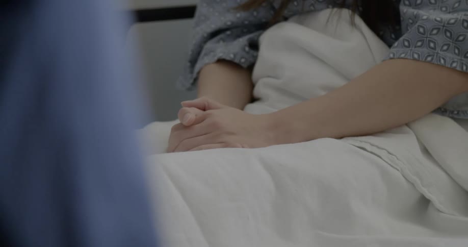 Patients hands are clasped on her lap while sitting in a hospital bed